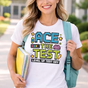 以下が含まれることがあります： ピクセル化された青と緑の文字で「ACE THE TEST」というテキストが入った白いTシャツ。シャツには「LEVEL UP TEST DAY」というテキストも入っています。その人はブルージーンズとティールバックパックを着用しています。