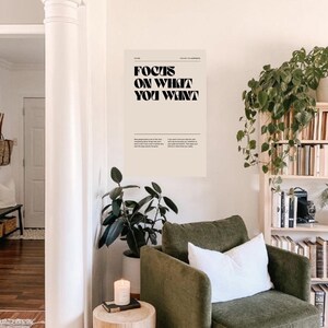 以下が含まれることがあります： 白いクッションのある緑色のアームチェアが、木のサイドテーブルと「欲しいものに集中しよう」と書かれたポスターのあるリビングルームに置かれている。