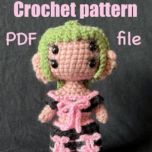 May include: Crochet pattern PDF file for a pink and black crocheted doll with green hair and large black eyes. The doll is wearing a pink and black outfit with bows.