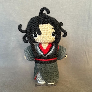 May include: Crocheted amigurumi doll with black hair, a red and black patterned robe, and a neutral-colored face. The doll is standing on a gray surface. The text "@venticrafts_official" is at the bottom of the image.