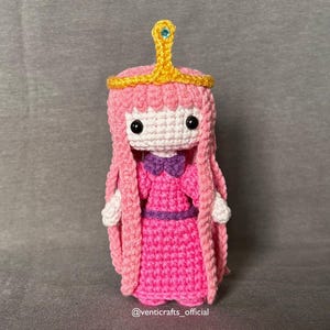 以下が含まれることがあります： アドベンチャー・タイムのプリンセス・バブルガムの手作りかぎ針編み人形。人形はピンクのドレス、長いピンクの髪、青い宝石が付いた金の王冠が特徴です。人形の高さは約15cmです。