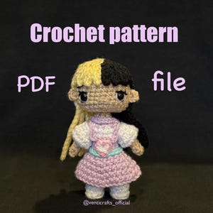 May include: Crochet pattern PDF file for a crocheted doll with black and yellow hair, wearing a purple dress with a pink heart on the front. The doll is standing on a black background.