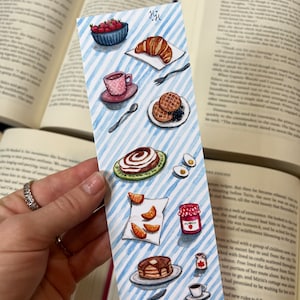 以下が含まれることがあります： 青と白のストライプの背景に、水彩画で描かれた朝食のブックマークのイラスト。ブックマークには、クロワッサン、イチゴのボウル、コーヒーカップ、ワッフルのプレート、シナモンロール、オレンジのスライスのプレート、ジャムの瓶、パンケーキの積み重ね、紅茶のカップなど、さまざまな朝食食品が描かれています。
