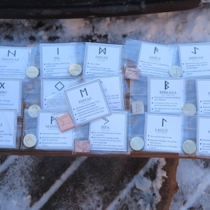 May include: A selection of rune sets, each in a clear plastic bag with a card detailing the rune's name and meaning. The runes are in shades of beige and copper. The cards are white with black text.
