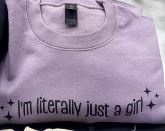 Puede incluir: Una sudadera de color lavanda con el texto "I'm literally just a girl" impreso en negro. La sudadera tiene una etiqueta negra con la palabra "MEDIUM" impresa en ella.