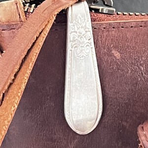 May include: A close-up of a brown leather bag with a silver metal zipper pull. The zipper pull is shaped like a spoon and has an ornate design.