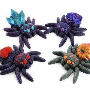 May include: Four black spider figurines with different colored accents. One spider has a blue crystal, one has a purple rose, one has a red heart, and one has an orange pumpkin.