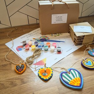 May include: A DIY craft kit to make your own decorative heart garland. The kit includes wooden hearts, paint, brushes, twine, and instructions. The hearts are painted in bright colours with various designs, including an eye, stripes, and flowers. The kit is packaged in a brown box with the words "Cultivate Create Handmade Kit" printed on it.
