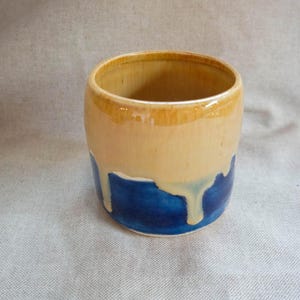 May include: A handmade ceramic cup with a cylindrical shape. The top and interior are a light tan colour, with a band of darker tan around the rim. The bottom half is a deep blue, with dripped tan glaze.