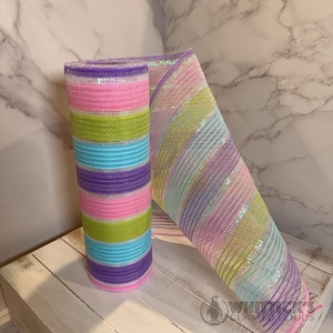May include: A roll of colorful deco mesh in shades of pink, green, blue, and purple. The mesh is striped and has a rolled-up section and a section that is unrolled. The mesh is on a white wooden surface.