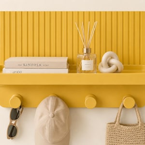 May include: A mustard yellow wall-mounted shelf with a vertical ribbed back panel and four circular hooks. The shelf holds two books titled The Kinfolk Home and at 100, a glass reed diffuser, and a beige decorative knot. Sunglasses, a cap, and a bag hang below.
