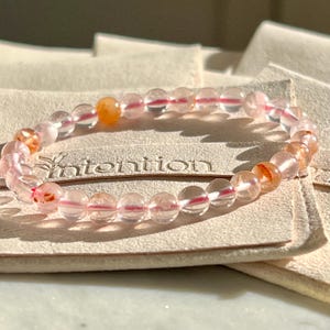 May include: A pink and orange crystal beaded bracelet with a pink thread running through the beads. The bracelet is on a light brown surface with the word "intention" printed on it.