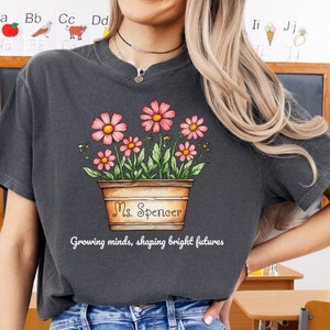 May include: A dark gray t-shirt with a flower pot design. The pot is brown with the text "Ms. Spencer" on it. The text "Growing minds, shaping bright futures" is below the pot.