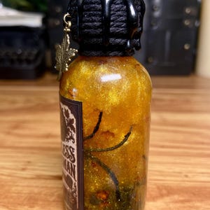Pode incluir: Uma garrafa de vidro transparente cheia de um l&iacute;quido dourado cintilante com elementos org&acirc;nicos escuros. O gargalo &eacute; envolto em cord&atilde;o preto com gotas de cera preta. Um pingente de folha de bordo cor de bronze pende do gargalo.