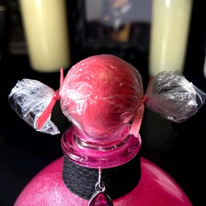 May include: A pink spherical object wrapped in clear plastic with red ribbon ties on both sides, sitting atop a pink bottle. A black fabric collar encircles the bottle neck, featuring a silver-toned pendant with a teardrop-shaped pink gem.