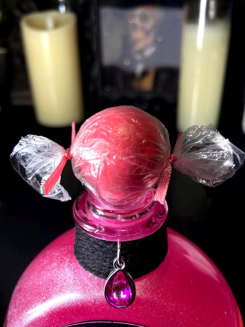 May include: A pink spherical object wrapped in clear plastic with red ribbon ties on both sides, sitting atop a pink bottle. A black fabric collar encircles the bottle neck, featuring a silver-toned pendant with a teardrop-shaped pink gem.