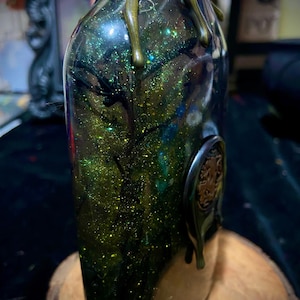 May include: A clear glass bottle filled with green liquid and gold glitter, featuring dark branch-like structures inside. The neck is wrapped in twine and copper-colored chain, with metallic green wax drips and a circular emblem on the side.