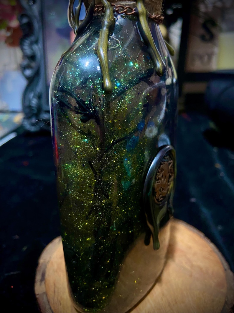 May include: A clear glass bottle filled with green liquid and gold glitter, featuring dark branch-like structures inside. The neck is wrapped in twine and copper-colored chain, with metallic green wax drips and a circular emblem on the side.