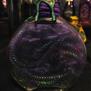 Puede incluir: Una botella de vidrio redonda llena de un l&iacute;quido arremolinado de color morado oscuro y negro con part&iacute;culas de purpurina verde iridiscente. El cuello de la botella est&aacute; envuelto en hilo morado y sellado con cera verde oscuro que gotea.