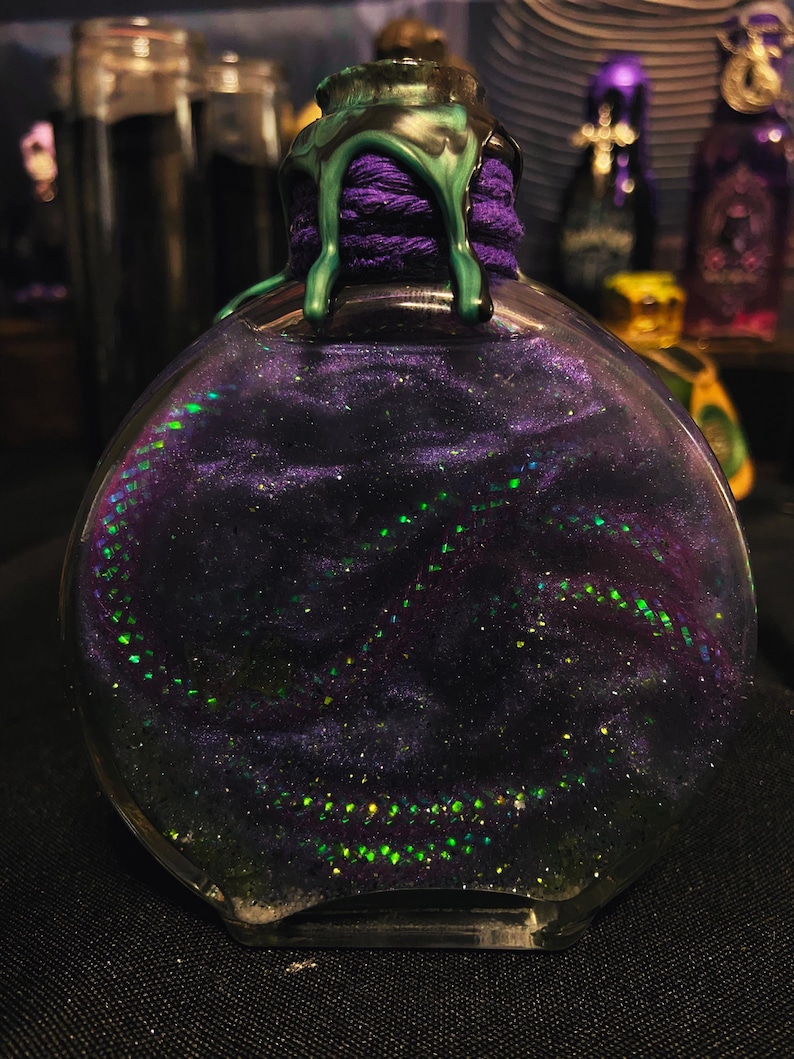 Puede incluir: Una botella de vidrio redonda llena de un l&iacute;quido arremolinado de color morado oscuro y negro con part&iacute;culas de purpurina verde iridiscente. El cuello de la botella est&aacute; envuelto en hilo morado y sellado con cera verde oscuro que gotea.