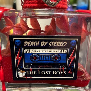 Puede incluir: Una botella de vidrio transparente llena de l&iacute;quido rojo y follaje rojo, con una etiqueta que muestra un gr&aacute;fico de casete azul, el texto DEATH BY STEREO, CRY LITTLE SISTER y THE LOST BOYS, rematada con cordel rojo y un dije de colmillos.