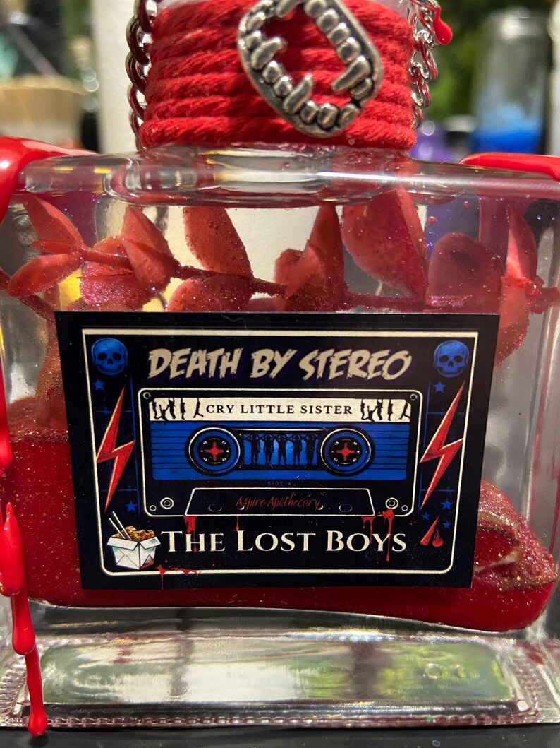 Puede incluir: Una botella de vidrio transparente llena de l&iacute;quido rojo y follaje rojo, con una etiqueta que muestra un gr&aacute;fico de casete azul, el texto DEATH BY STEREO, CRY LITTLE SISTER y THE LOST BOYS, rematada con cordel rojo y un dije de colmillos.