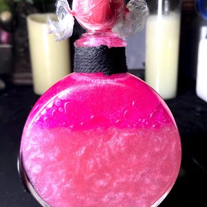May include: A round glass bottle filled with a vibrant pink, shimmering liquid. The neck of the bottle is wrapped in black twine, and the top is sealed with a pink sphere wrapped in clear plastic, creating a candy-like appearance.