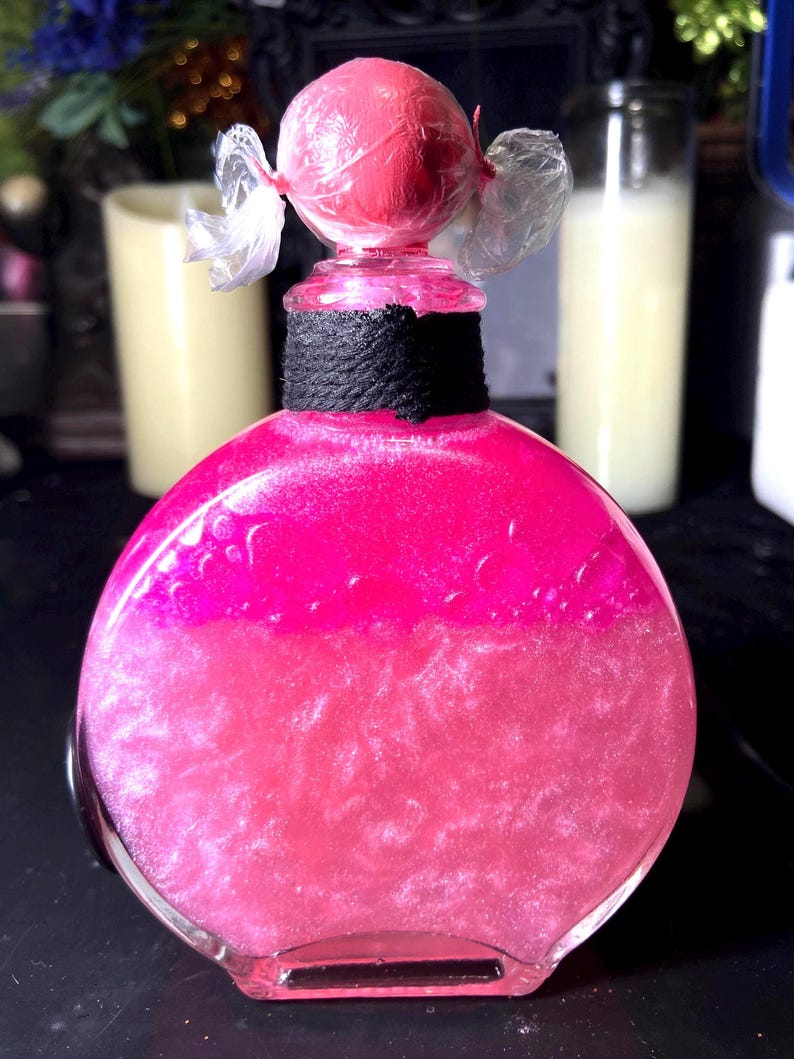 May include: A round glass bottle filled with a vibrant pink, shimmering liquid. The neck of the bottle is wrapped in black twine, and the top is sealed with a pink sphere wrapped in clear plastic, creating a candy-like appearance.