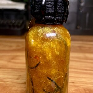 Pode incluir: Uma pequena garrafa de vidro transparente cheia de um l&iacute;quido dourado cintilante contendo elementos bot&acirc;nicos verde-escuros e vermelhos. O gargalo &eacute; envolto em cord&atilde;o preto e selado com cera preta brilhante que escorre pelas laterais.