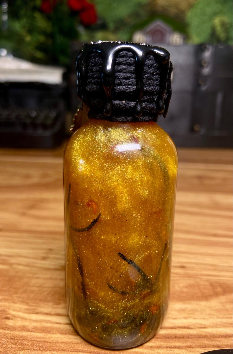 Pode incluir: Uma pequena garrafa de vidro transparente cheia de um l&iacute;quido dourado cintilante contendo elementos bot&acirc;nicos verde-escuros e vermelhos. O gargalo &eacute; envolto em cord&atilde;o preto e selado com cera preta brilhante que escorre pelas laterais.