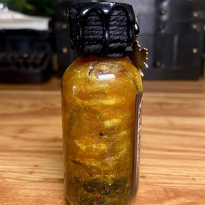 Pode incluir: Uma garrafa de vidro transparente cheia de um l&iacute;quido dourado cintilante e part&iacute;culas escuras. O gargalo da garrafa &eacute; envolto em barbante preto e selado com cera preta. Um pequeno pingente de folha de bordo dourada pende do gargalo e um r&oacute;tulo retangular est&aacute; fixado na lateral.