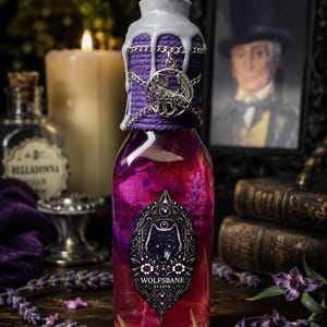 May include: A glass bottle filled with pink and purple liquid, featuring a label with a wolf illustration and the text "WOLFSBANE ELIXIR". The neck is wrapped in purple twine and silver chains, topped with a silver wolf charm and grey wax.