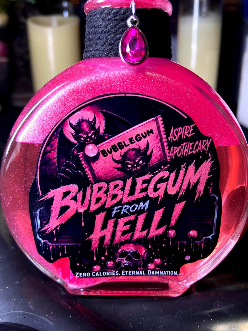 May include: A round glass bottle filled with shimmering pink liquid, featuring a black label with the text BUBBLEGUM FROM HELL, ASPIRE APOTHECARY, and ZERO CALORIES. ETERNAL DAMNATION. The bottle neck is wrapped in black cord with a pink teardrop charm.