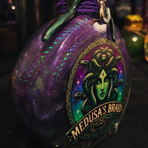 Puede incluir: Una botella de vidrio llena de l&iacute;quido morado con purpurina, con una etiqueta que muestra una ilustraci&oacute;n de Medusa de piel verde y el texto Medusa's Braid. El cuello est&aacute; envuelto en cordel morado y sellado con cera verde y un dije de cristal.