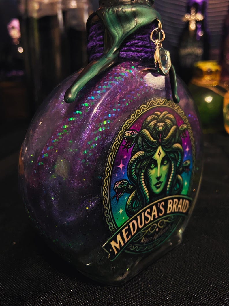 Puede incluir: Una botella de vidrio llena de l&iacute;quido morado con purpurina, con una etiqueta que muestra una ilustraci&oacute;n de Medusa de piel verde y el texto Medusa's Braid. El cuello est&aacute; envuelto en cordel morado y sellado con cera verde y un dije de cristal.