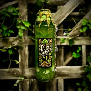 May include: A green glass bottle with gold accents and a label that reads "Fairy Garden" with a green glitter inside. The bottle is hanging from a wooden fence.