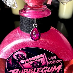 May include: A pink glittery liquid in a glass bottle with a black wrapped neck and a silver teardrop charm. The label features a devil illustration and the text BUBBLEGUM FROM HELL! and ASPIRE APOTHECARY. The bottle is topped with a clear plastic wrapper.