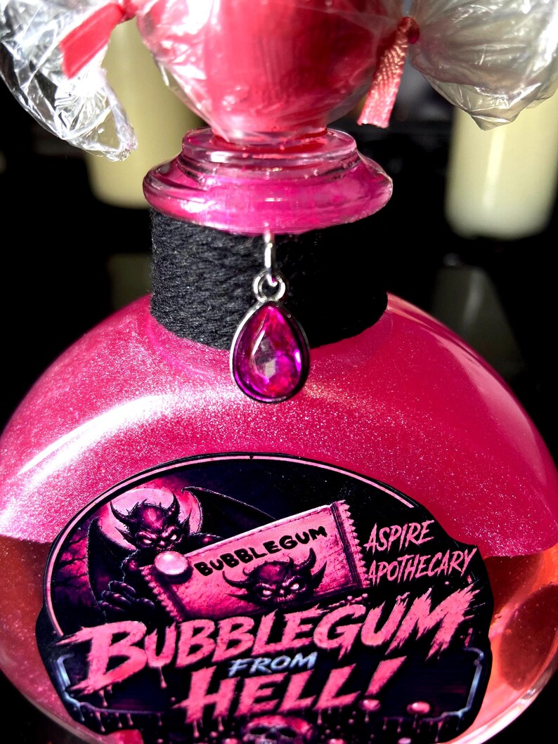 May include: A pink glittery liquid in a glass bottle with a black wrapped neck and a silver teardrop charm. The label features a devil illustration and the text BUBBLEGUM FROM HELL! and ASPIRE APOTHECARY. The bottle is topped with a clear plastic wrapper.