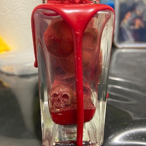 Puede incluir: Una botella de vidrio transparente llena de objetos rojos brillantes con forma de calavera y l&iacute;quido rojo. El cuello de la botella est&aacute; envuelto en cuerda roja y tiene una cadena de metal plateado. Cera roja espesa gotea por el exterior del vidrio.