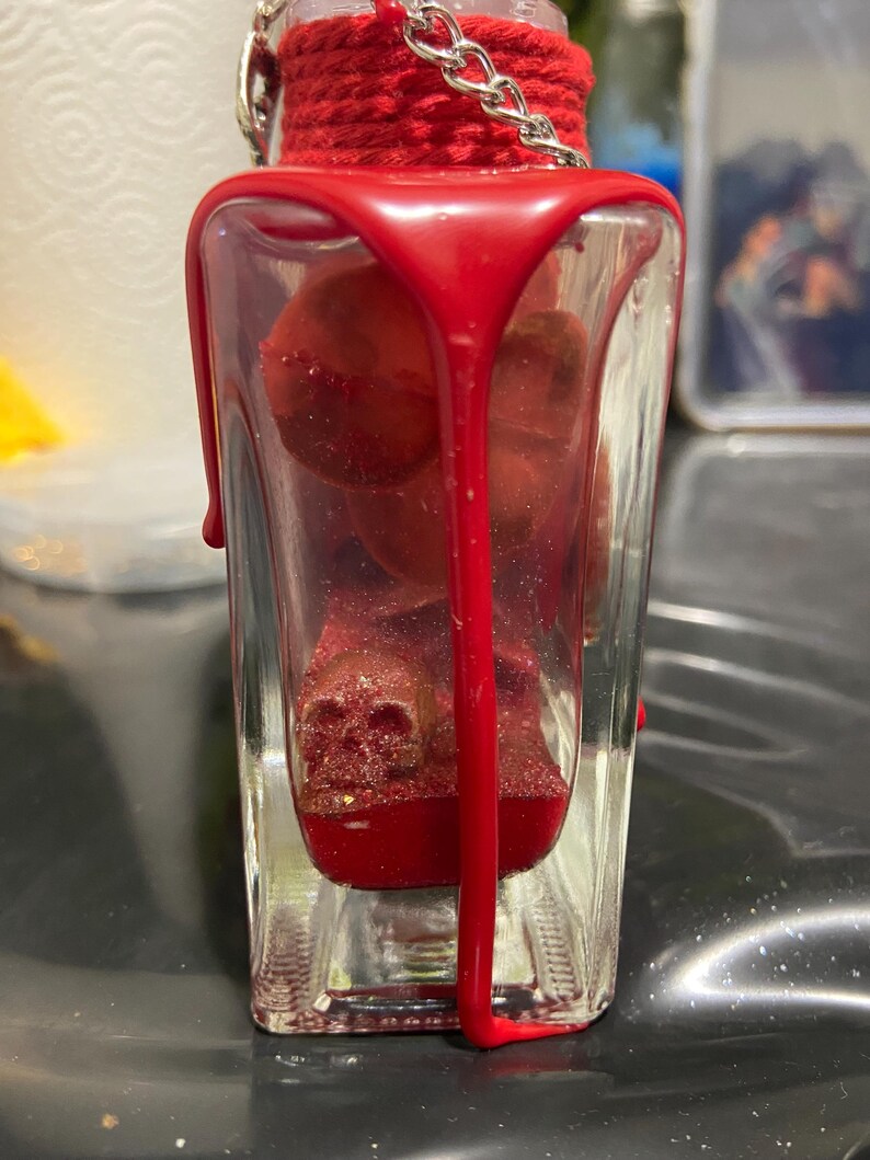 Puede incluir: Una botella de vidrio transparente llena de objetos rojos brillantes con forma de calavera y l&iacute;quido rojo. El cuello de la botella est&aacute; envuelto en cuerda roja y tiene una cadena de metal plateado. Cera roja espesa gotea por el exterior del vidrio.