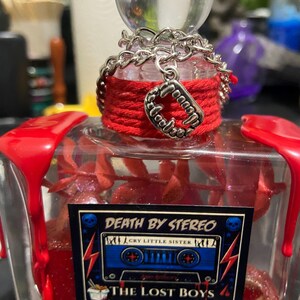 Puede incluir: Una botella de vidrio transparente con cera roja goteando desde las esquinas superiores. El cuello est&aacute; envuelto en hilo rojo con una cadena plateada y un dije de colmillos de vampiro. Una etiqueta rectangular muestra un casete azul y el texto DEATH BY STEREO.