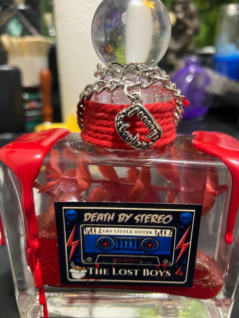Puede incluir: Una botella de vidrio transparente con cera roja goteando desde las esquinas superiores. El cuello est&aacute; envuelto en hilo rojo con una cadena plateada y un dije de colmillos de vampiro. Una etiqueta rectangular muestra un casete azul y el texto DEATH BY STEREO.