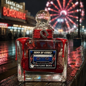 Puede incluir: Un frasco de perfume rectangular de cristal lleno de l&iacute;quido rojo, con gotas de cera roja en las esquinas superiores. La etiqueta dice DEATH BY STEREO, CRY LITTLE SISTER y THE LOST BOYS. Una cadena plateada y un dije de colmillos de vampiro rodean el cuello.