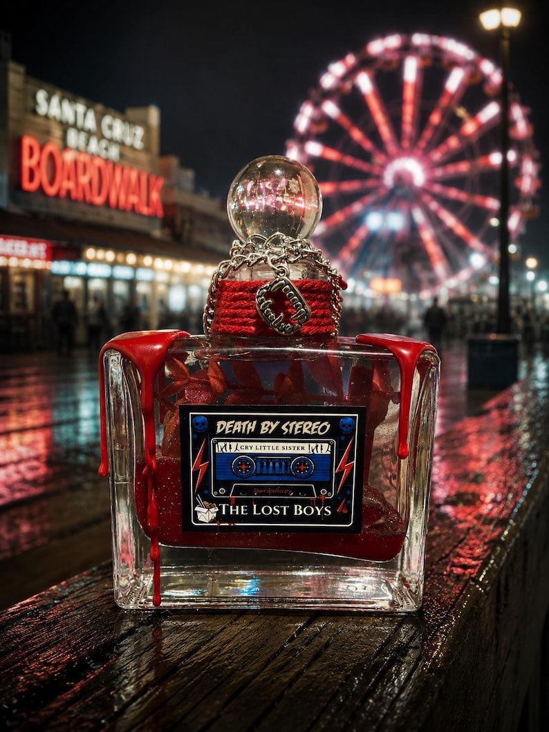Puede incluir: Un frasco de perfume rectangular de cristal lleno de l&iacute;quido rojo, con gotas de cera roja en las esquinas superiores. La etiqueta dice DEATH BY STEREO, CRY LITTLE SISTER y THE LOST BOYS. Una cadena plateada y un dije de colmillos de vampiro rodean el cuello.