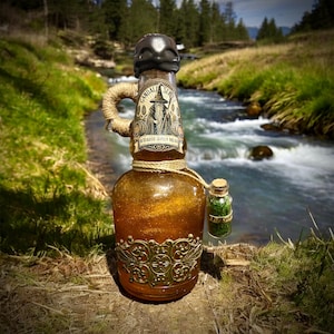 May include: A decorative glass bottle with a dark skull-shaped stopper, a label that reads "Gandalf's Mead", and a small attached vial labeled "Pipeweed". The bottle has a gold-colored liquid and is adorned with a gold filigree design.