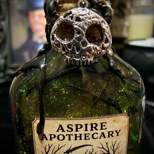 May include: A green glass bottle filled with glittery liquid, featuring a silver skull charm attached to the neck. Black wax drips down from the top. A label reads ASPIRE APOTHECARY with an illustration of a headless horseman riding a horse.