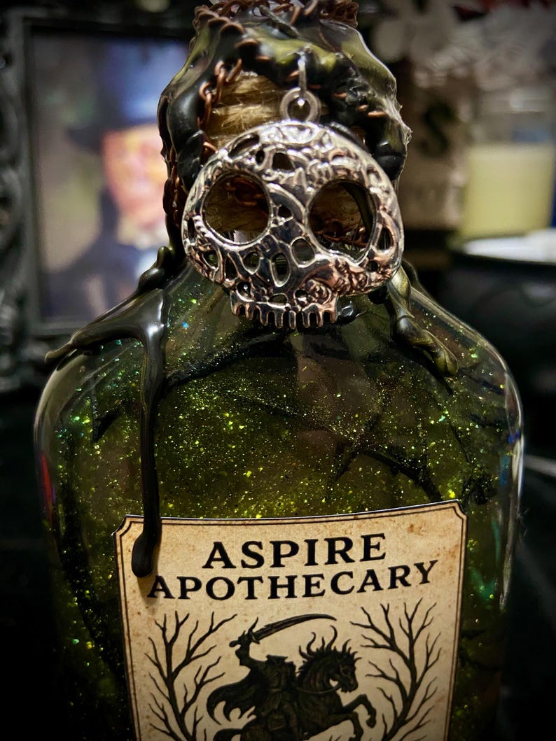 May include: A green glass bottle filled with glittery liquid, featuring a silver skull charm attached to the neck. Black wax drips down from the top. A label reads ASPIRE APOTHECARY with an illustration of a headless horseman riding a horse.
