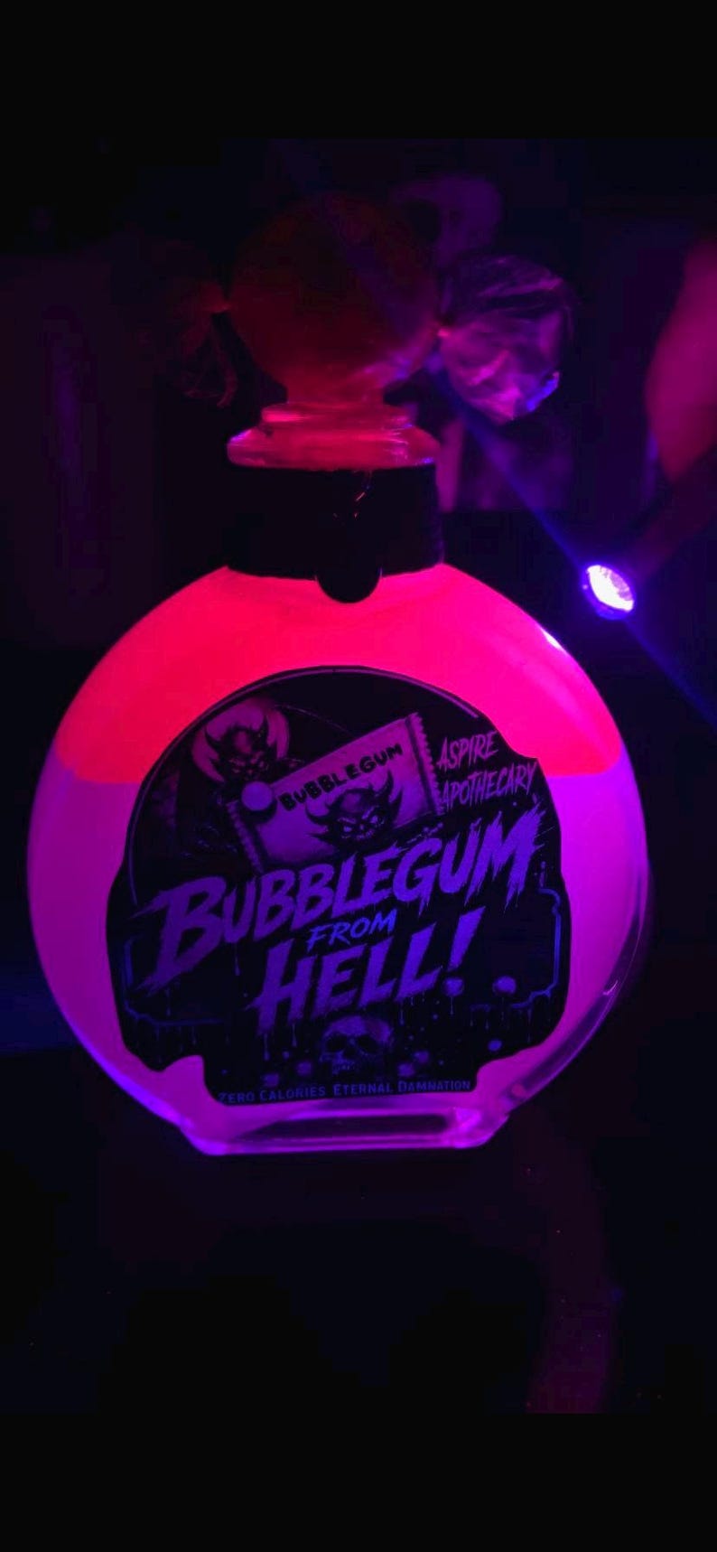 May include: A round glass bottle filled with a glowing neon pink liquid under ultraviolet light. The label features a black graphic with a devil illustration and the text BUBBLEGUM FROM HELL, ASPIRE APOTHECARY, and ZERO CALORIES ETERNAL DAMNATION.