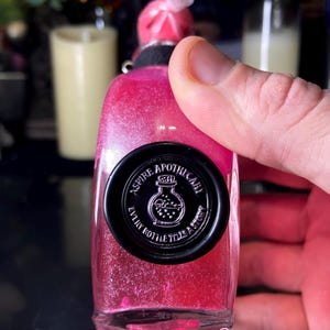 May include: A clear glass bottle filled with vibrant pink glittery liquid, topped with a pink wax seal. A circular black wax seal on the front features the text ASPIRE APOTHECARY and EVERY BOTTLE TELLS A STORY surrounding a small bottle icon.