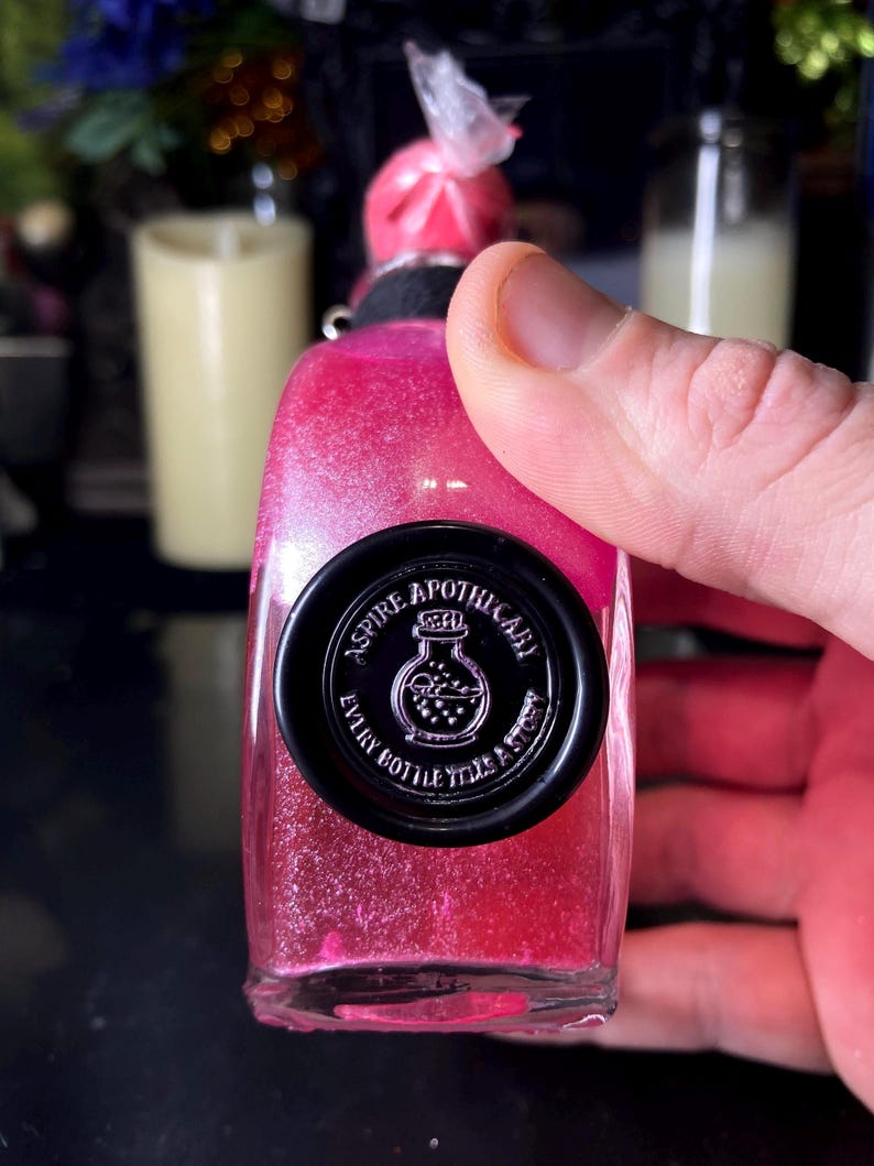 May include: A clear glass bottle filled with vibrant pink glittery liquid, topped with a pink wax seal. A circular black wax seal on the front features the text ASPIRE APOTHECARY and EVERY BOTTLE TELLS A STORY surrounding a small bottle icon.
